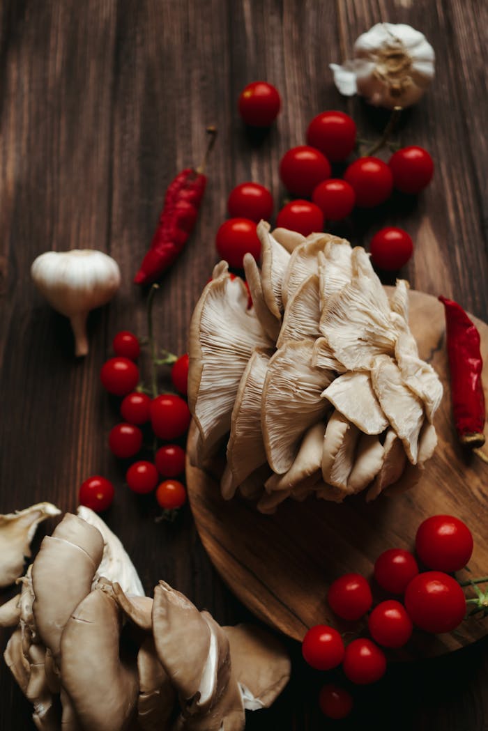 A rustic display of oyster mushrooms, cherry tomatoes, garlic, and chili peppers on a wooden surface.